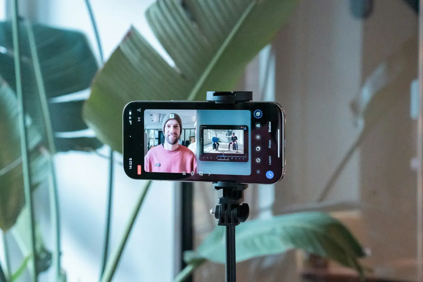 A smartphone on a tripod, its screen showing the man it is recording in a pink shirt and grey beanie, with plants behind
